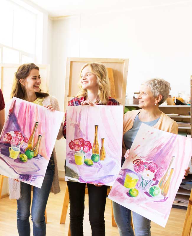 Three women holding their bright paintings and looking at each other.