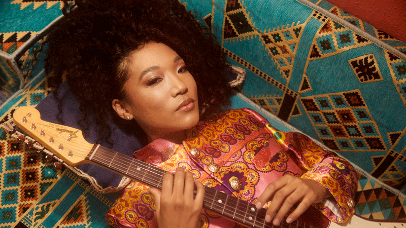 A woman laying on a colorful blanket with a guitar.